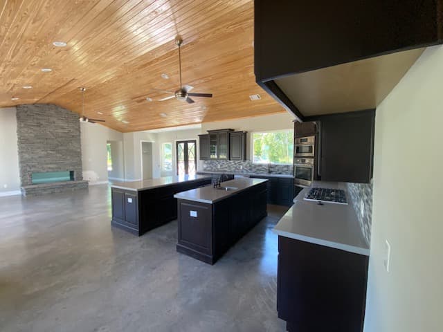 Modern kitchen featuring two dark islands, wood-paneled vaulted ceiling, and a large stone fireplace.