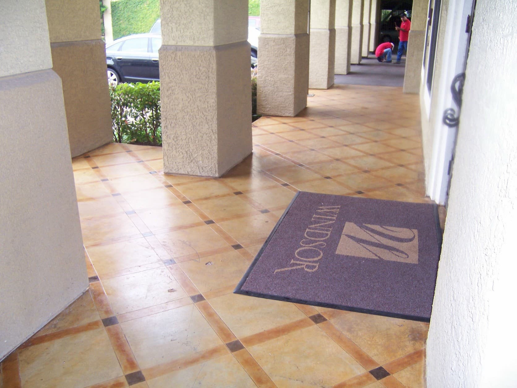 Covered walkway with large pillars, patterned tiles, and a purple Windsor doormat near workers.